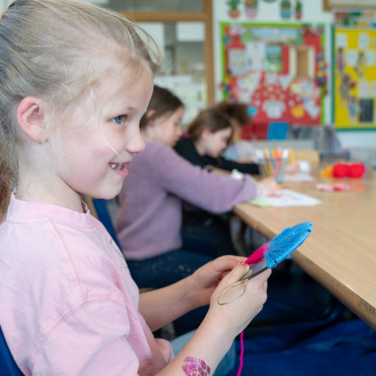Young girl in a pink shirt crafting with yarn and feathers at a classroom table, with other students and colorful boards in the background.