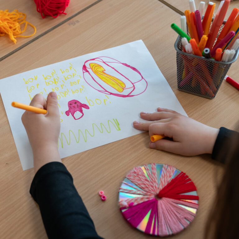 Child draws on a white sheet with yellow text and pink-yellow shapes while colorful markers sit in a cup nearby on a wooden table.