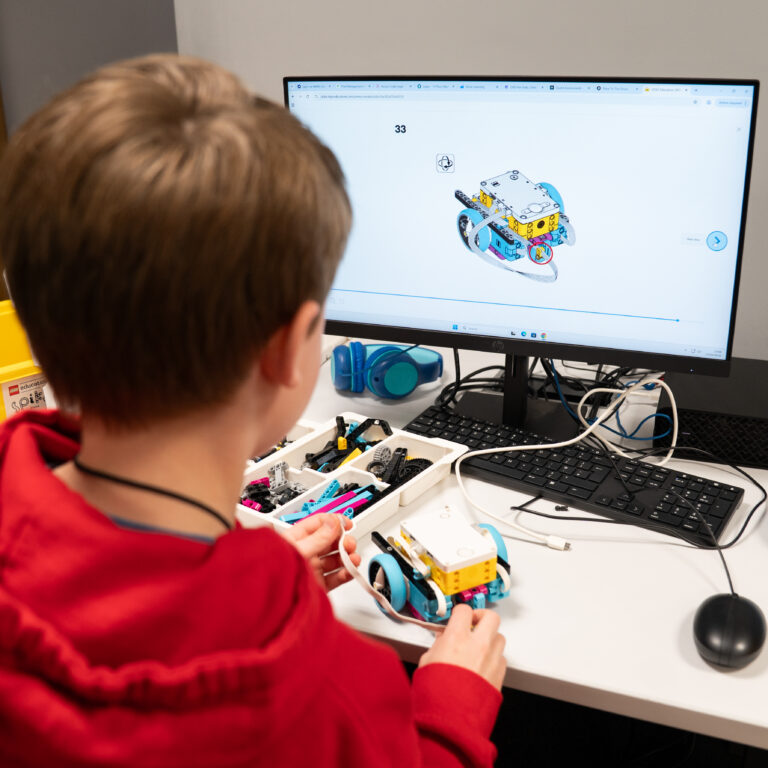 Child in a red jacket building a LEGO robotics model at a desk, with a computer screen showing a robot design in the background