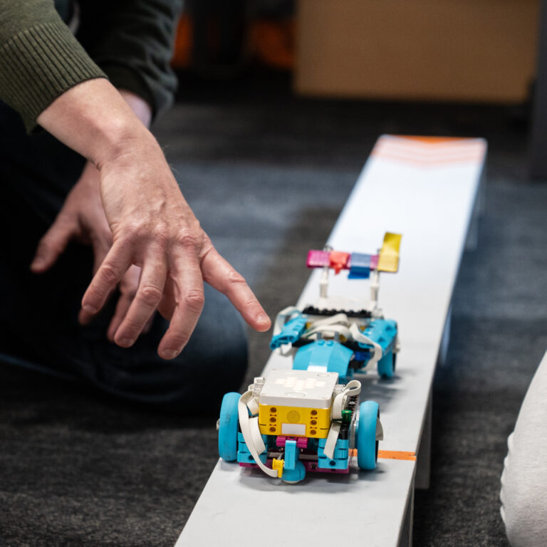 Hands adjust a colorful LEGO robot on a white bench while two people sit nearby, engaging with the project.