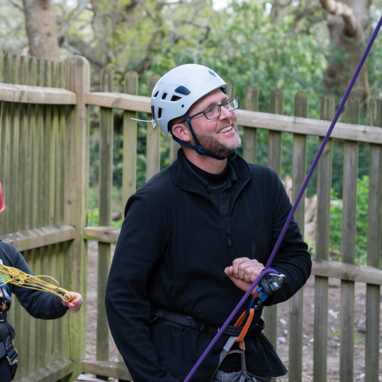 Man and child wearing helmets and harnesses outdoors, preparing with ropes for a climbing activity by a wooden fence.