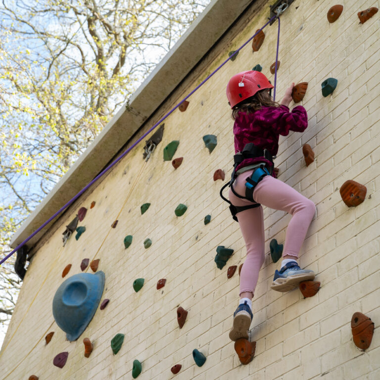 Child wearing a red helmet climbs an outdoor rock wall with colorful holds, secured by a harness and rope.