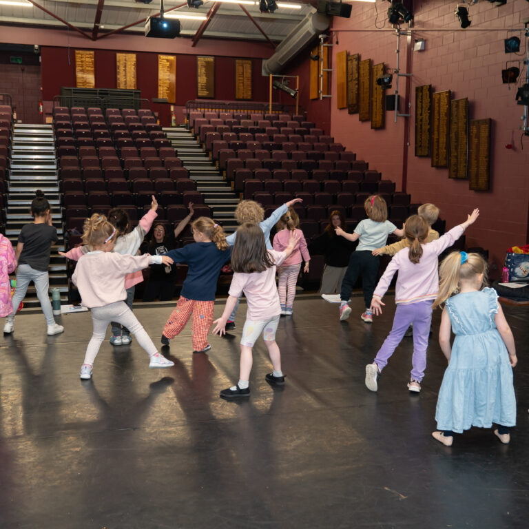 Group of children dancing on a theater stage with raised arms, maroon auditorium seats in the background.
