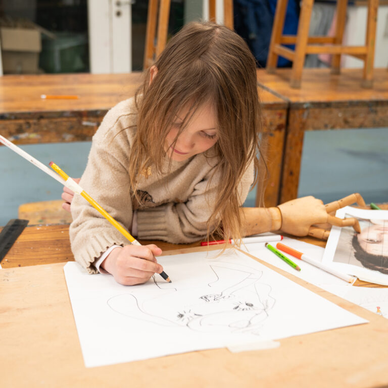 Young girl with long hair sketching on papers at a cluttered art table in a studio setting.