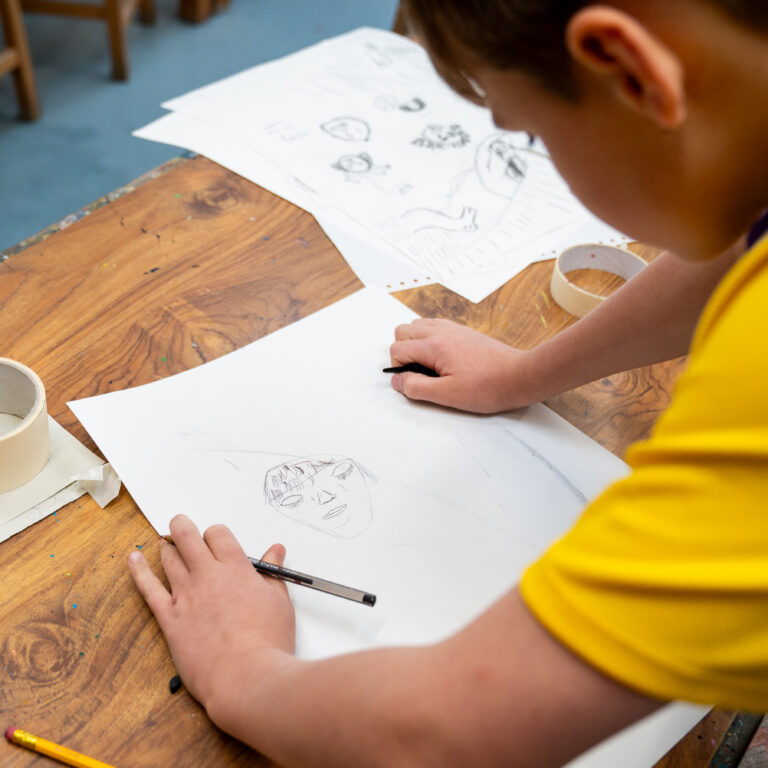 Young artist drawing on white paper at a wooden art table, with tape rolls and pencils nearby.