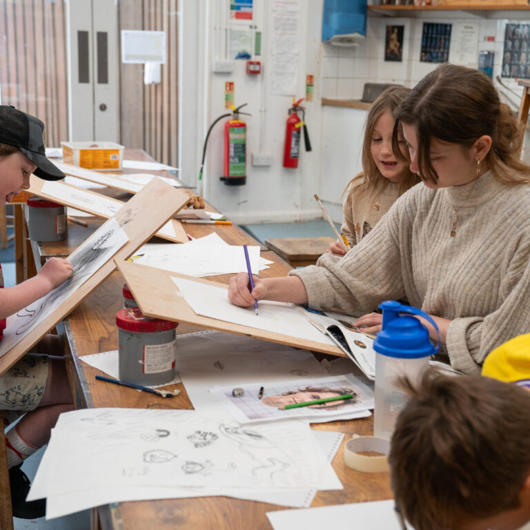 Kids at a long wooden table drawing and sketching on paper in a bright art studio.