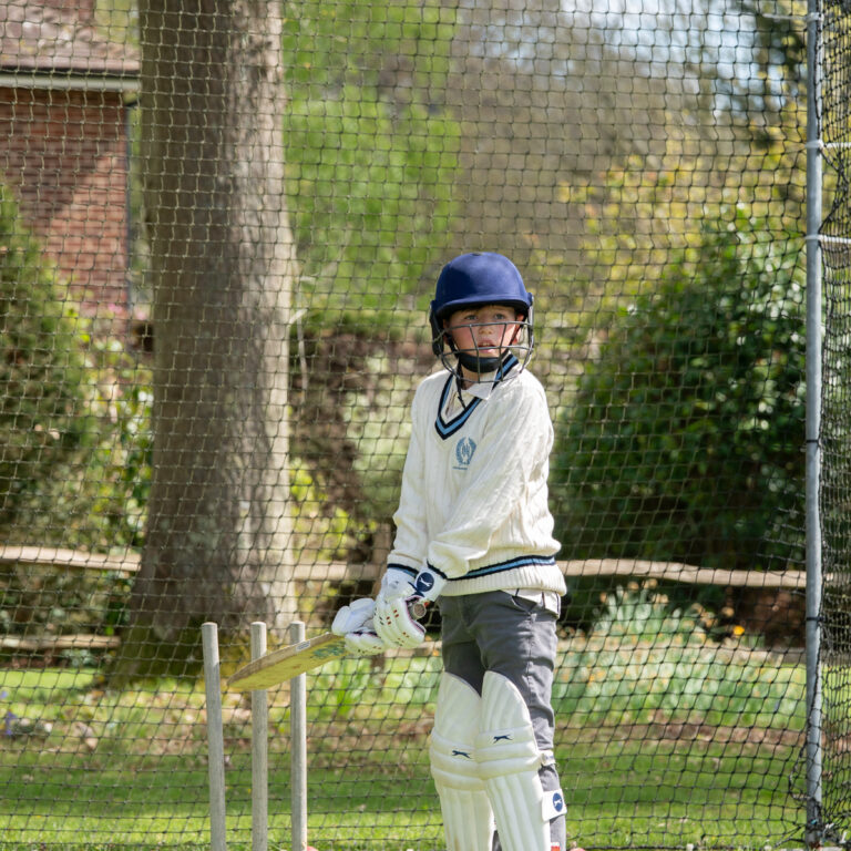 Cricket player wearing helmet and pads stands near the wickets behind a protective net in a practice area.