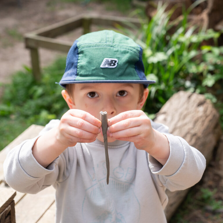 Child wearing a green-and-blue hat shows a small twig toward the camera outdoors on a wooden deck.