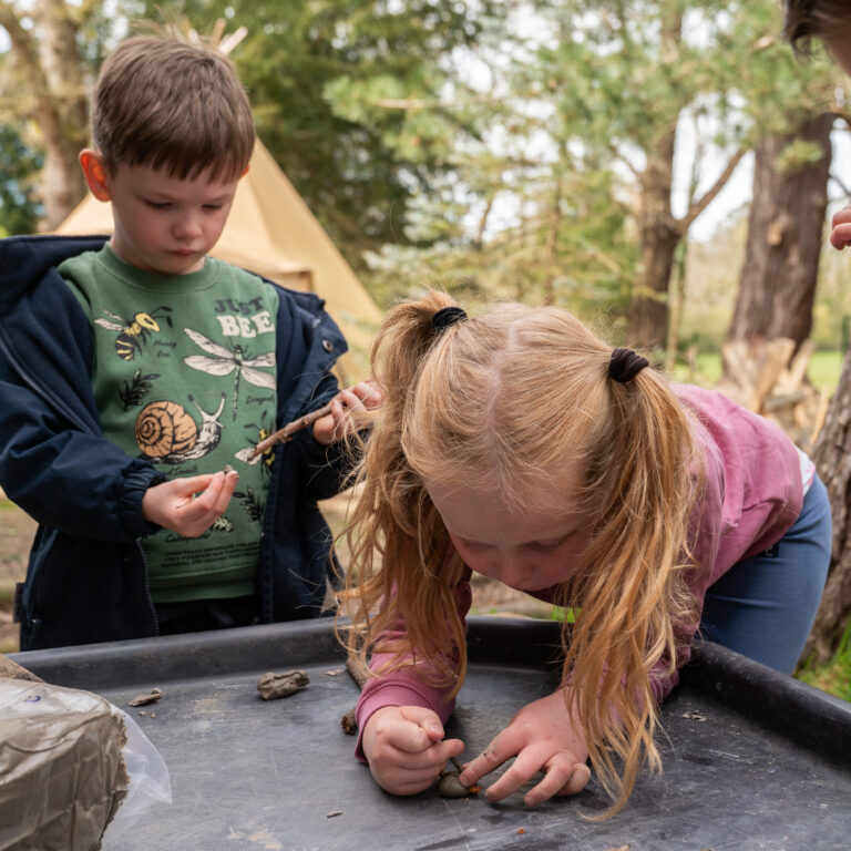 Children outdoors at a clay crafting table, shaping small clay pieces with hands and sticks, supervised by others nearby in a park setting.