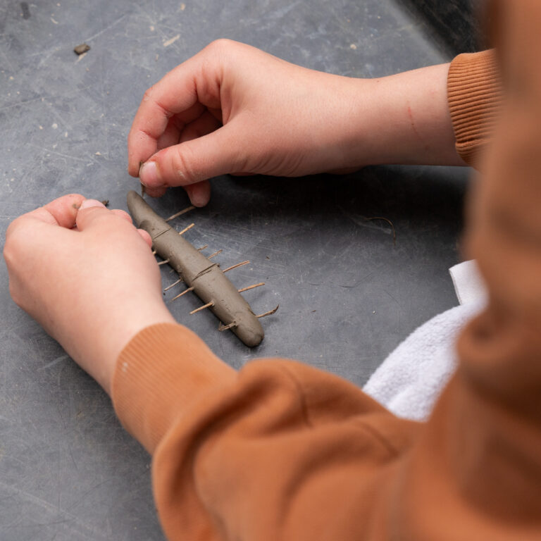 Close-up of hands shaping and smoothing a clay coil on a dark work surface, with a rough, textured clay piece nearby.