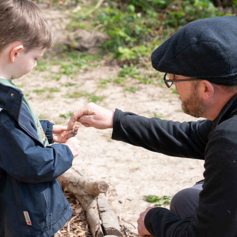 Adult man with a dark cap kneels beside a young boy outdoors, handing him a small object between them.