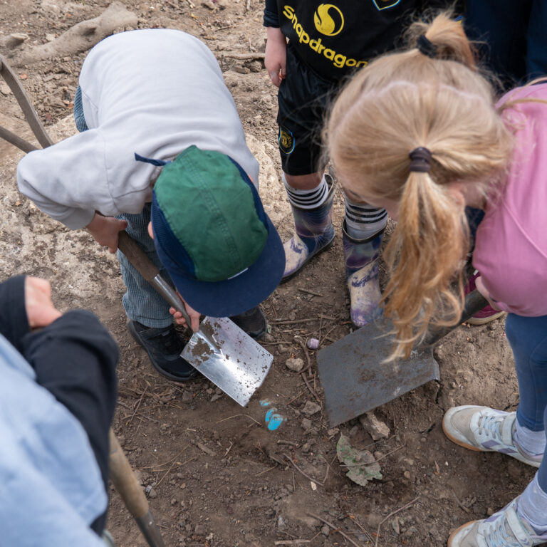 Group of children outdoors, gathered around as they dig in dirt with small shovels.