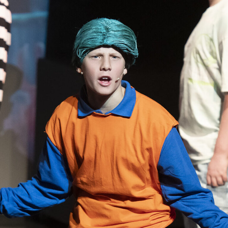 Young performer with teal hair in an orange and blue costume on stage, arms outstretched, with other cast members in the background.